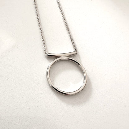 A silver-colored circle necklace with a rolo-style chain and a pendant featuring a circle with a curved line above it.