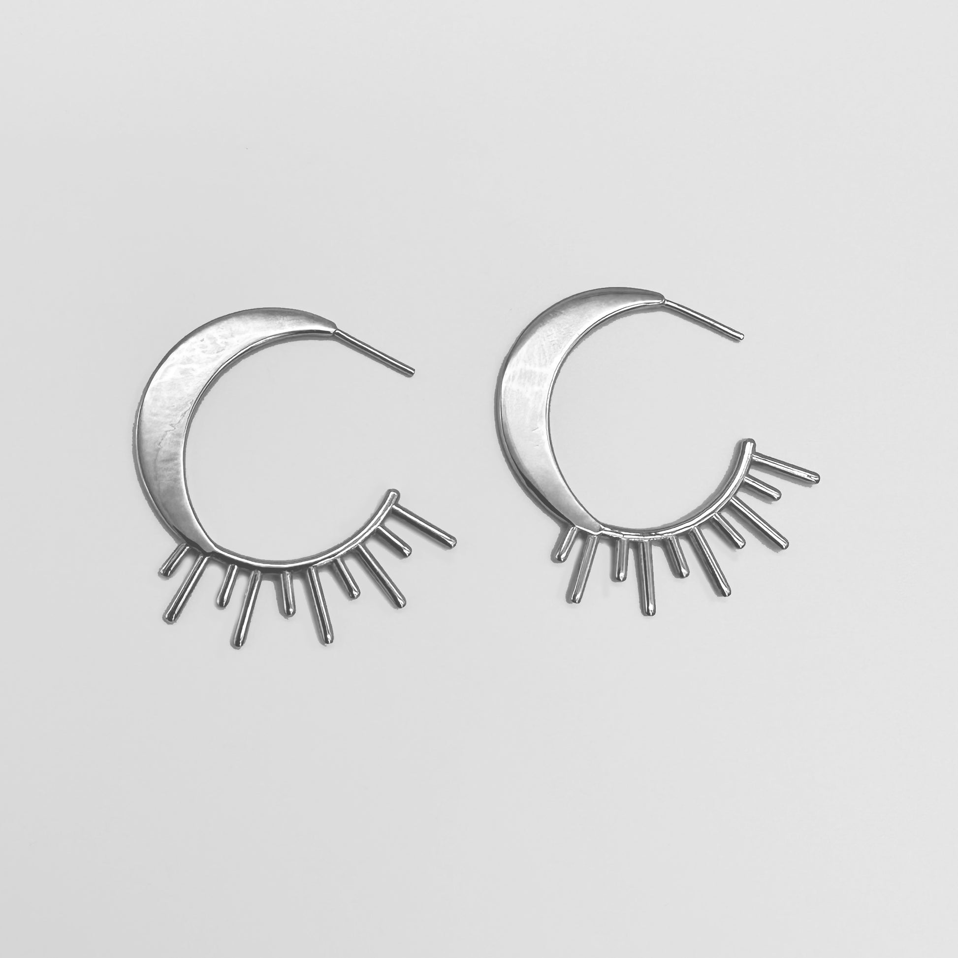 A pair of silver-colored celestial hoop earrings designed with rays resembling sunbeams extending from the hoop, resembling a sun and moon pattern.