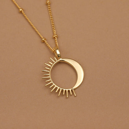 A gold necklace featuring a crescent moon and sun pendant, displayed on a solid warm brown background.