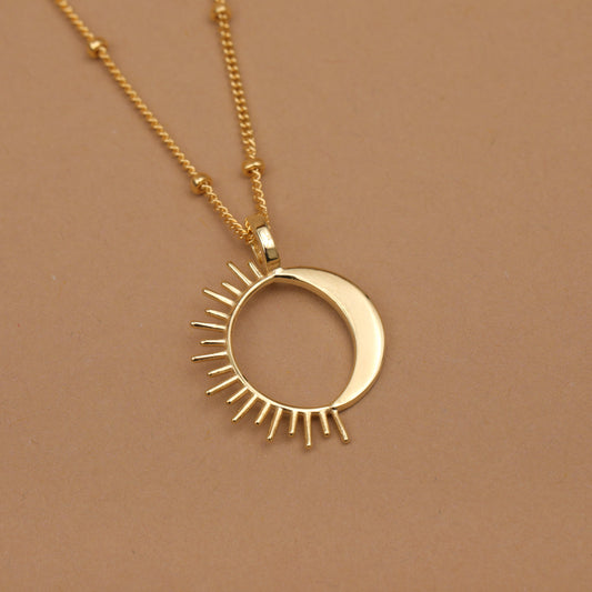 A gold necklace featuring a crescent moon and sun pendant, displayed on a solid warm brown background.