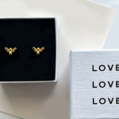 Gold bee-shaped earrings in a black box with 'LOVE' card on a white surface