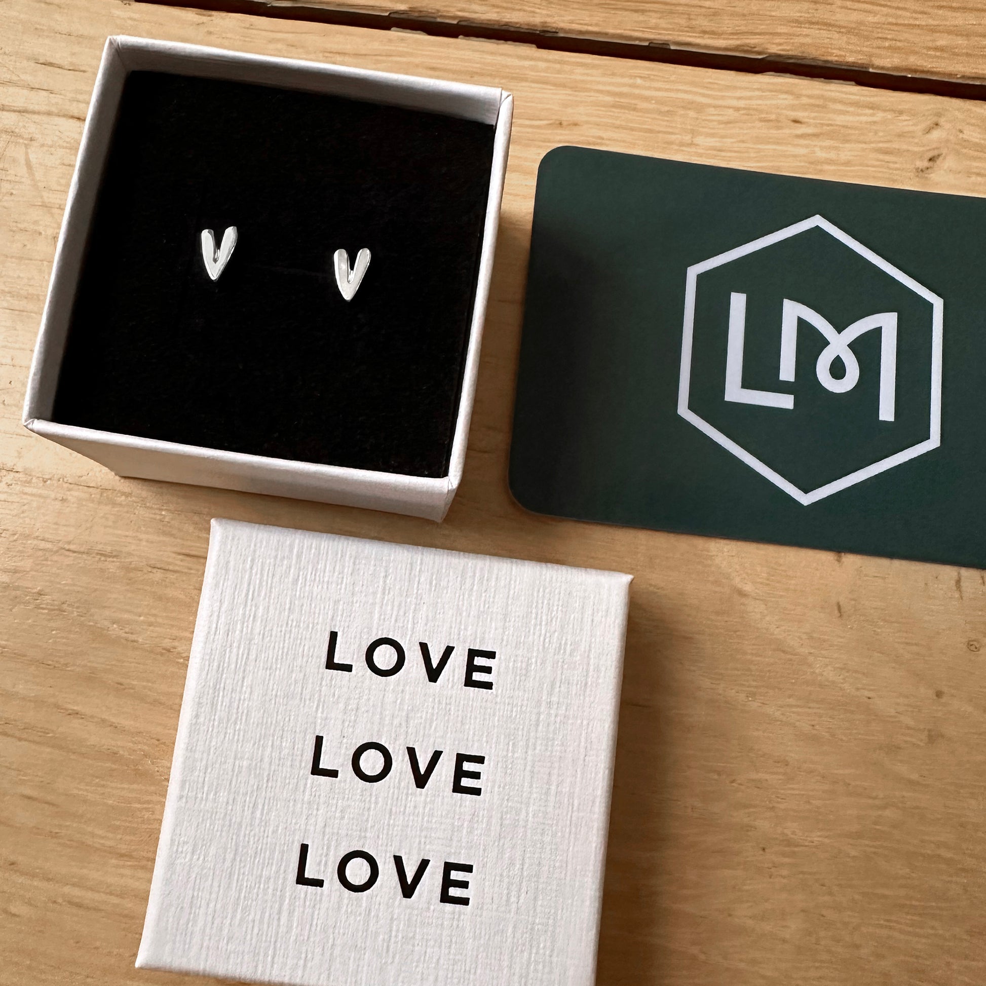 Heart-shaped earrings in a box with 'LOVE' card and LM brand card on a wooden surface