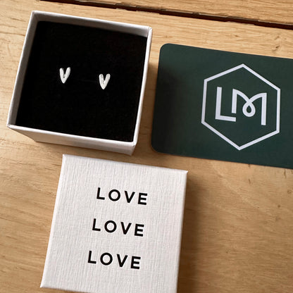 Heart-shaped earrings in a box with 'LOVE' card and LM brand card on a wooden surface