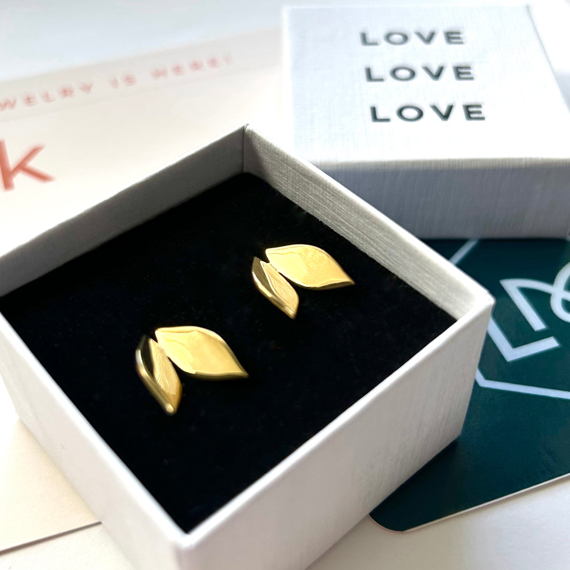 Gold leaf-shaped earrings in a box with 'LOVE' text in the background