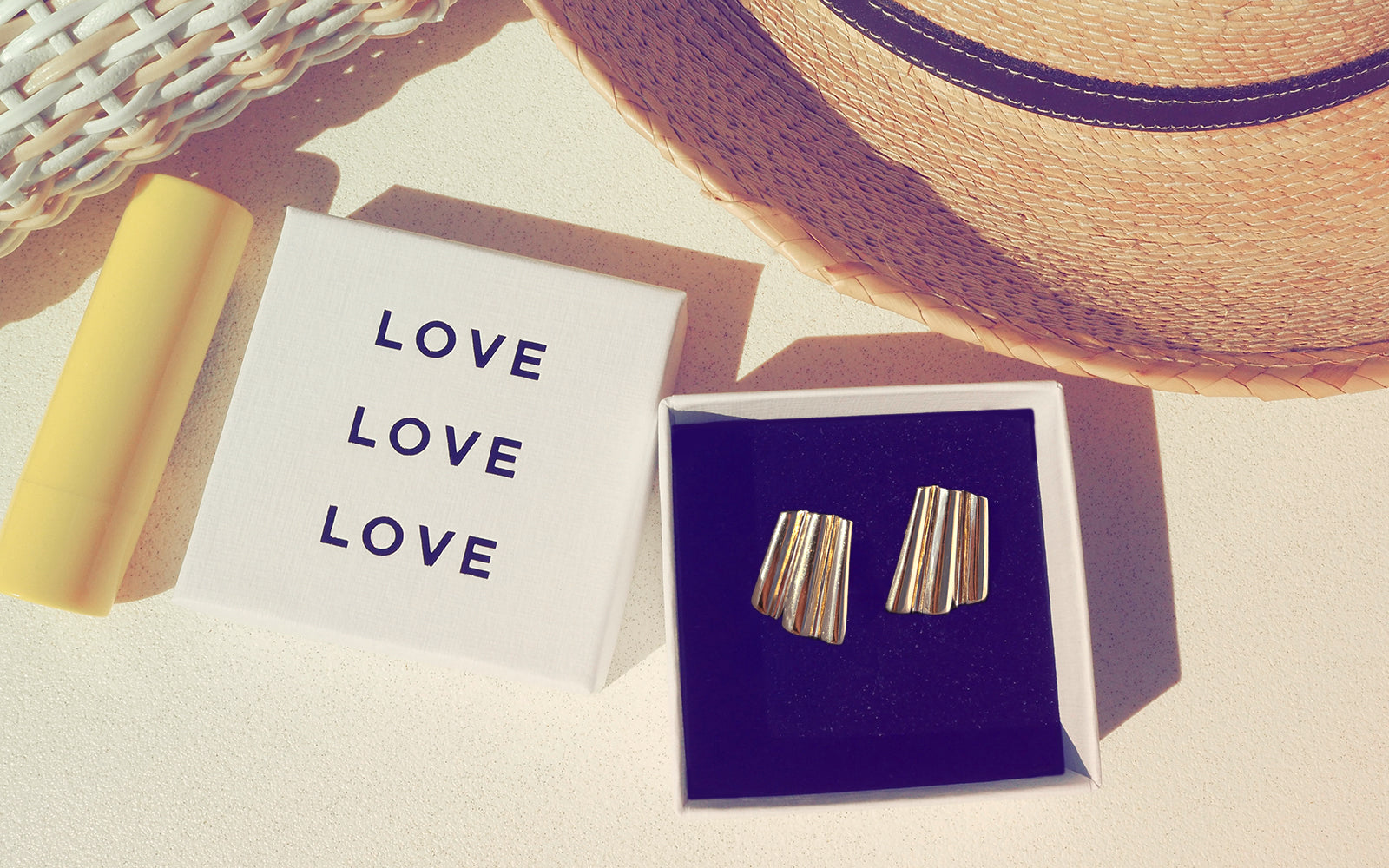 Gold earrings in a box with 'LOVE' card and straw hat on light background