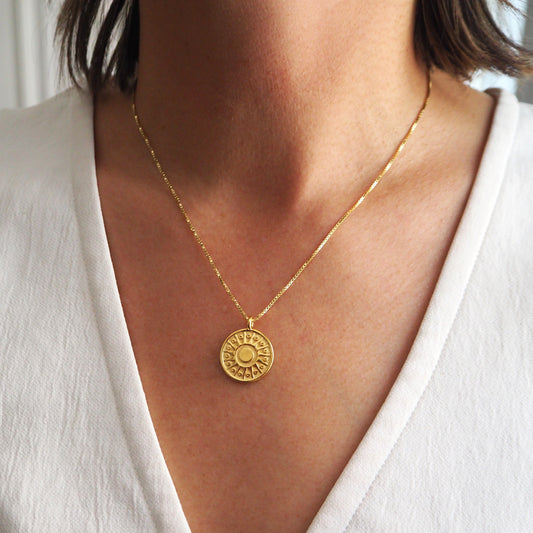 Gold necklace with a circular pendant worn by a person in a white outfit.