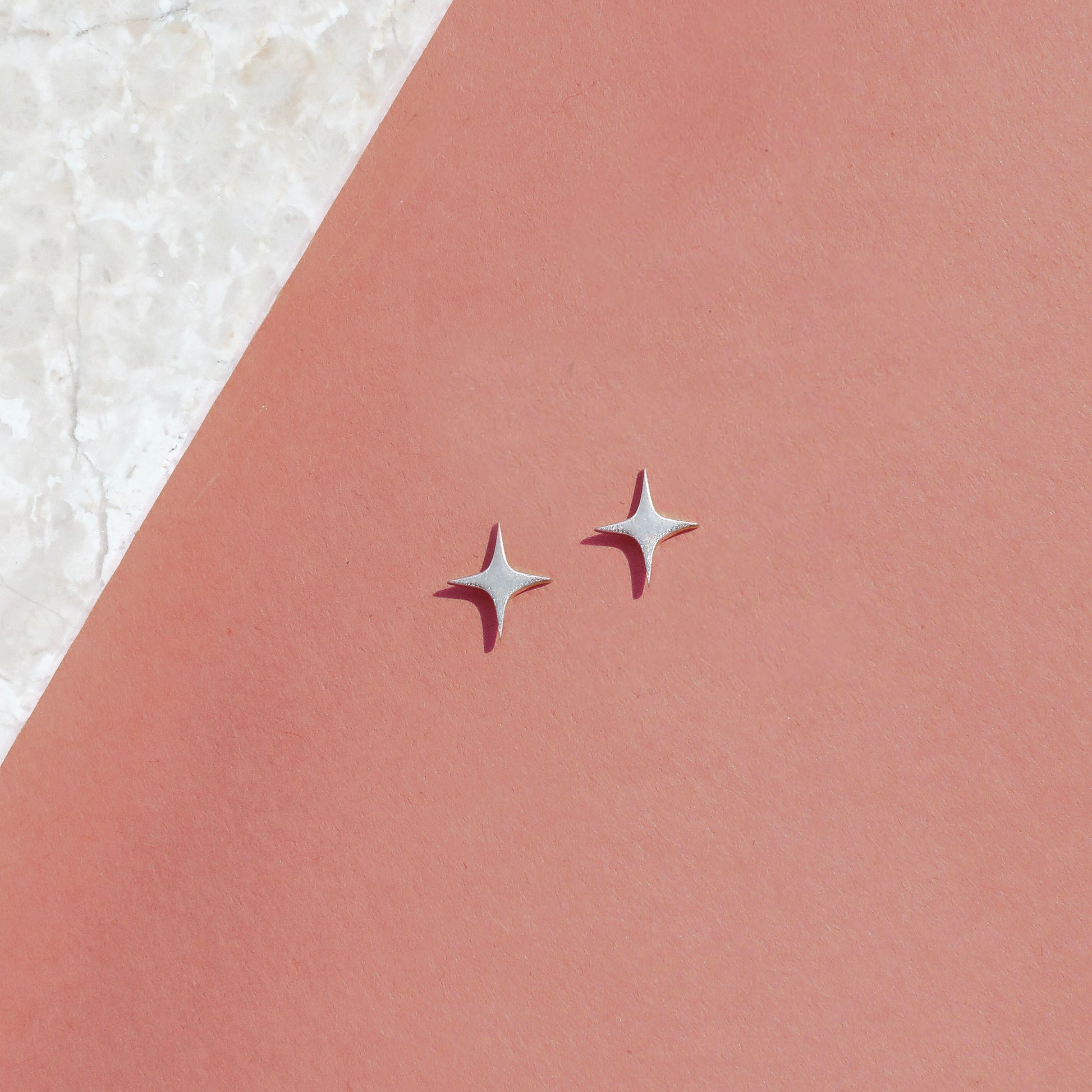 A pair of silver-colored star-shaped stud earrings against a pink background.