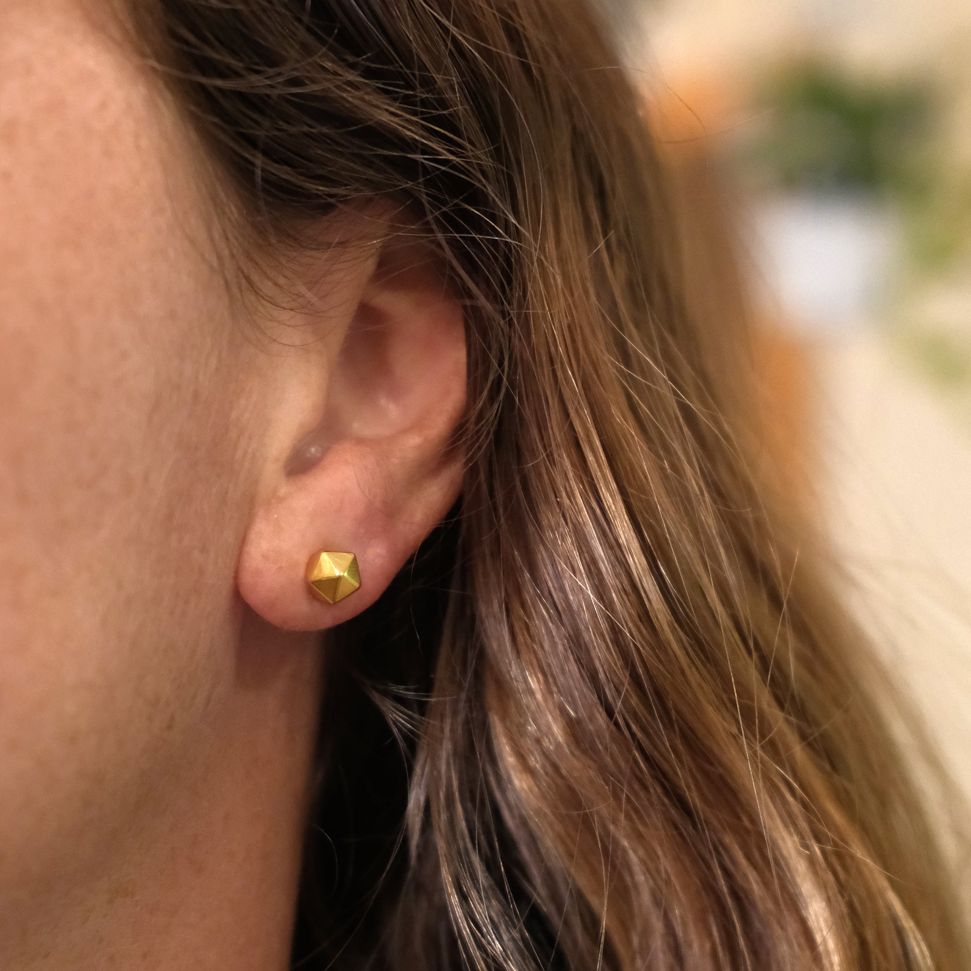 Close-up of an ear wearing a gold hexagonal earring with blurred background